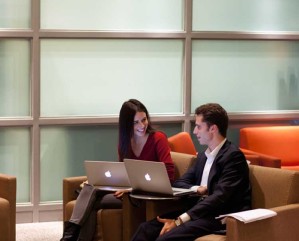 A couple of people are seated on comfortable chairs with open laptops in a modern office setting, engaged in a conversation with smiles.