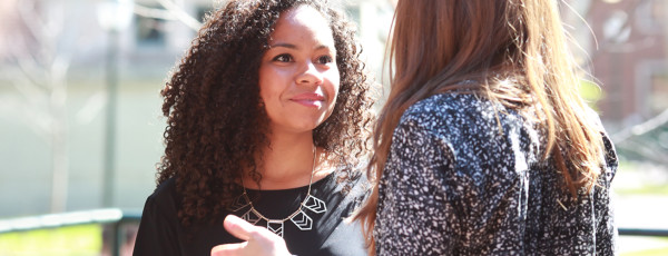 Two people are having a conversation outdoors. One person is facing away from the camera, and the other person is smiling slightly. The scene is well-lit, suggesting a sunny day.