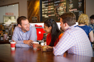 A small group of people sitting at a table in a cafe, engaged in conversation. They are holding coffee cups, with a menu board visible in the background.