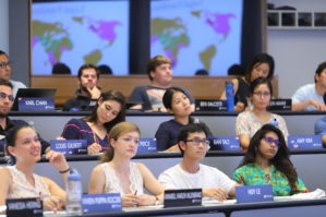 A classroom setting with several students seated, taking notes, and listening to a lecture. Name plaques with students'' names and flags are visible. A world map is projected on a screen in the background.