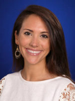 conversation_Brent Headshot of a person with long brown hair, wearing a white garment and large earrings, smiling against a blue background.