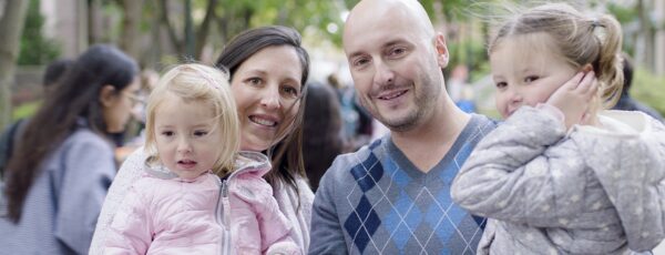 A smiling family of four outdoors, consisting of two adults and two children, surrounded by trees and people in the background.