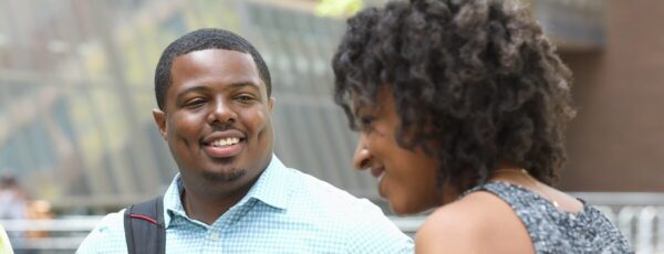 Two people are engaged in conversation outdoors, smiling at each other. The atmosphere appears casual and friendly.