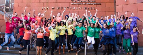 A diverse group of people is energetically jumping in front of a building marked "Jon M. Huntsman Hall" at The Wharton School, wearing matching colorful T-shirts.