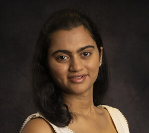 Headshot of a person with long dark hair wearing a light-colored top, set against a dark background.