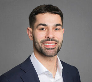 Aboneaaj_Rakan_final Headshot of a person with short dark hair and a beard, wearing a dark suit jacket and white shirt, smiling in front of a gray background.