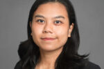 Headshot of a person with long dark hair, wearing a dark blazer, set against a plain gray background.