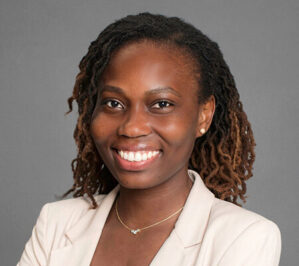 Headshot of a person smiling, wearing a light-colored blazer and a necklace, with curly hair styled in locs, against a gray background.