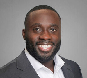 Ahoua, Wilfred_final Headshot of a person in a suit and white shirt, smiling against a gray background.