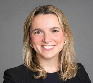 Headshot of a smiling person with long, light hair wearing a dark top, set against a neutral background.