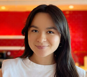 Chulack, Anna_final Headshot of a person with long dark hair, wearing a white shirt, smiling in front of a blurred red background.