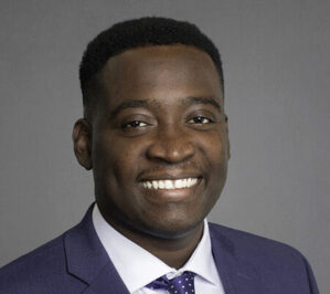 Cobbinah, James_final Headshot of a person wearing a dark blue suit, light shirt, and polka dot tie, smiling in front of a plain gray background.
