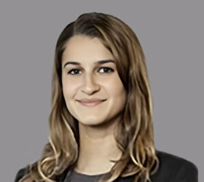 Economos, Ioana_finals Headshot of a person with long brown hair, smiling, against a gray background.