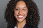 A headshot of a person with curly hair smiling, wearing a dark blazer over a light top, against a plain background.