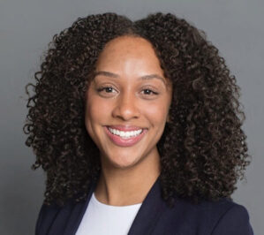 Geter, Elyshia_final A headshot of a person with curly hair smiling, wearing a dark blazer over a light top, against a plain background.