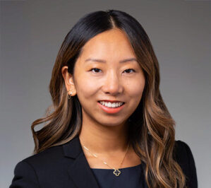 Lindy Gou Headshot of a person with long, wavy hair wearing a black jacket and a necklace, smiling against a neutral background.