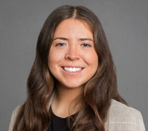 Handler, Miara_final Headshot of a person with long, dark hair, wearing a light-colored blazer and a black shirt, smiling against a gray background.