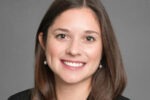 Headshot of a person with long brown hair, smiling, wearing pearl earrings and a dark blazer, set against a plain gray background.