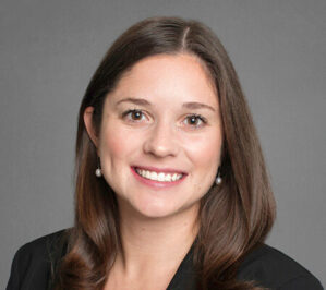 Healy, Sarah_final Headshot of a person with long brown hair, smiling, wearing pearl earrings and a dark blazer, set against a plain gray background.