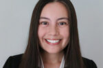 Headshot of a smiling person with long, straight hair, wearing a black blazer and a white shirt against a neutral background.