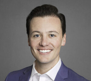 Hobbs, Christian Troy_final A headshot of a smiling person in a blue blazer and white shirt, set against a neutral gray background.