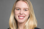 A headshot of a person with long, blond hair, smiling, and wearing a dark blazer against a gray background.