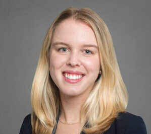 Jakubowicz, Addison Karlyse_final A headshot of a person with long, blond hair, smiling, and wearing a dark blazer against a gray background.