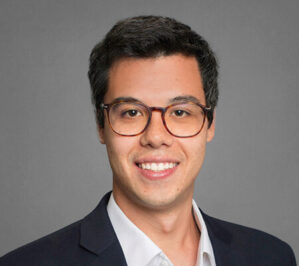 Jukemura, Pedro Kenzo_final Headshot of a person with short dark hair wearing glasses, a dark suit jacket, and a white shirt, smiling against a gray background.