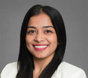 Kambli, Aditi_final Headshot of a person with long dark hair, smiling, wearing a white suit jacket against a plain background.