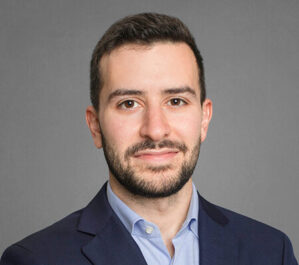 Kazan, Mark_final Headshot of a person with short dark hair and a beard, wearing a dark suit jacket and a light blue shirt, against a gray background.