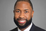 A professional headshot of a person with short hair and a beard, wearing a suit and tie, smiling against a neutral background.