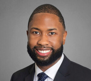 Lassiter, Quintin Delmart Jr._final A professional headshot of a person with short hair and a beard, wearing a suit and tie, smiling against a neutral background.