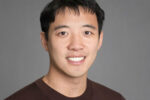 Headshot of a person with short, dark hair, wearing a brown sweater, smiling in front of a neutral gray background.