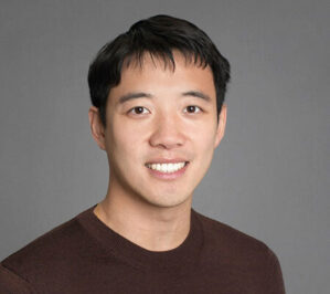 Headshot of a person with short, dark hair, wearing a brown sweater, smiling in front of a neutral gray background.