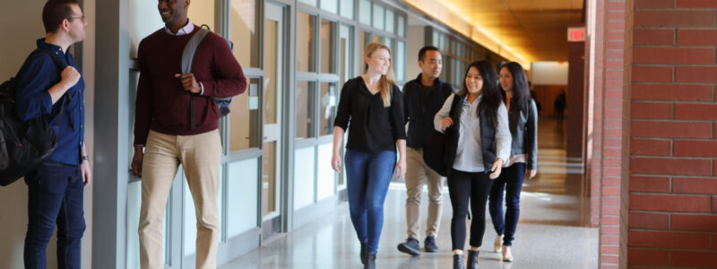 A group of people in a school or university hallway. Two are standing and talking on the left, while several others walk together on the right. They appear to be students, suggesting a learning environment.