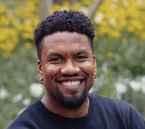 McDaniel, Samuel_final A person with short curly hair and a beard is smiling outdoors, wearing a dark shirt with a background of leafy trees.