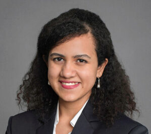 Mitra, Tiyasha_final Headshot of a person with curly dark hair, wearing a dark suit and white shirt, smiling against a neutral background.