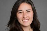 Headshot of a person with long dark hair smiling, wearing a dark blazer, set against a plain gray background.