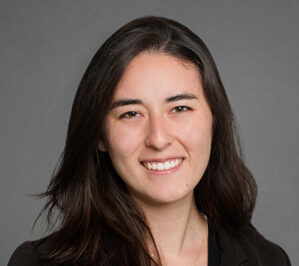 Nakamura Chapuis, Carolina_final Headshot of a person with long dark hair smiling, wearing a dark blazer, set against a plain gray background.