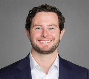 Nolan, Riley Charleston_final Headshot of a person with short, dark curly hair, wearing a white shirt and dark blazer, smiling at the camera.