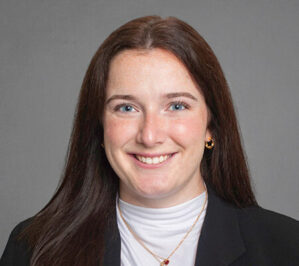 Odierno, Sabrina Wentworth_final Headshot of a person with long brown hair, wearing a black blazer over a white top, smiling against a gray background.