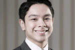 Headshot of a person in a suit and tie, smiling, with a blurred neutral background.