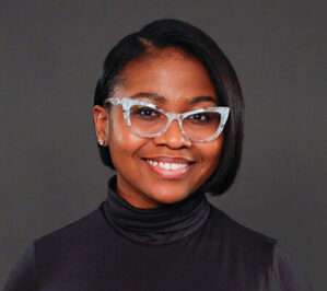 Plowright, Brianna Elizabeth_final Headshot of a person wearing glasses and a black turtleneck, smiling against a dark background.