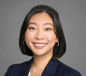 Rusznak, Michelle Fang_final Headshot of a person with short dark hair, smiling, wearing hoop earrings and a dark blazer against a plain background.
