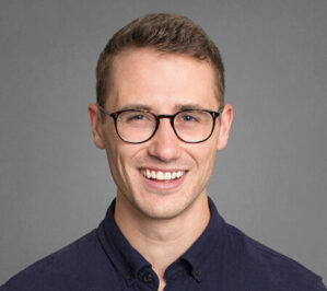 Seck, Matthew (Matt)_final A headshot of a person wearing glasses and a dark shirt, smiling against a plain background.