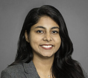 Sharma, Daksha_final Headshot of a person with long dark hair, wearing a gray blazer, smiling against a plain background.
