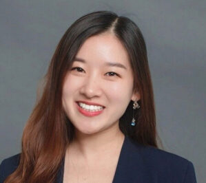 A headshot of a person smiling, wearing a dark blazer and earrings, with long hair, against a plain background.