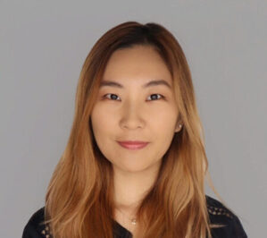 Headshot of a person with long, light brown hair and a neutral expression, wearing a dark top, set against a plain background.