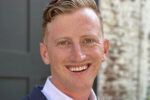 Headshot of a person smiling, wearing a suit with a white shirt. The background is slightly blurred, featuring a wall with mixed textures.