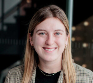 Headshot of a smiling person with long hair wearing a checkered blazer and dark shirt, standing indoors with a blurred background.